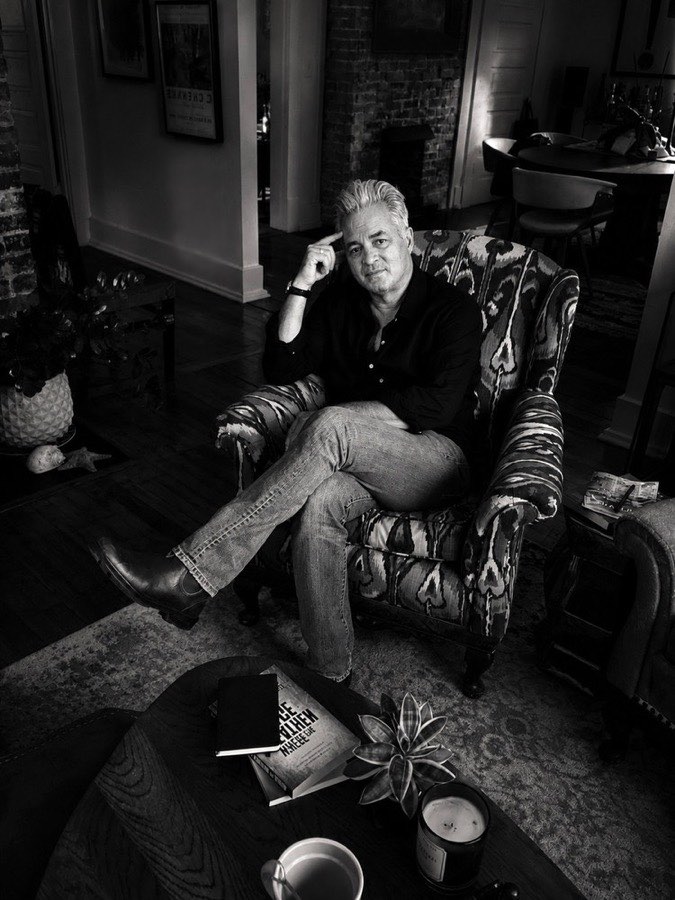 Stacy Dean Campbell seated in a patterned wing chair at home, photographed in black and white, a copy of the novel on the table in front of him.